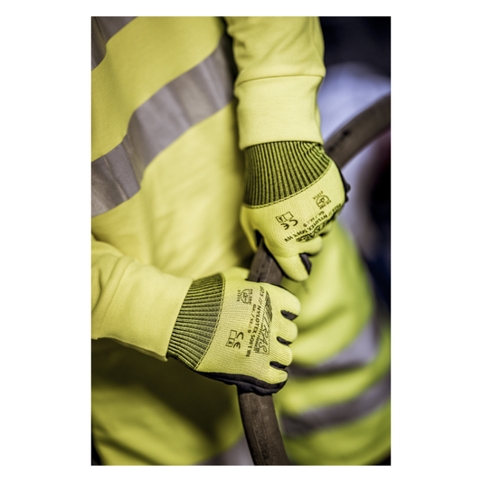 Eine Person, die eine gelbe Warnweste und Nylonhandschuhe NITRAS NYLOTEX SOFT HV von AS Arbeitsschutz GmbH, neongelb, teilbeschichtet – mit schwarzem Text und Sicherheitssymbolen – trägt, hält ein dickes, dunkles Kabel oder einen Schlauch.