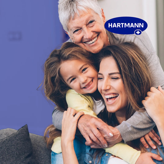 Drei Generationen von Frauen - eine Seniorin, eine Erwachsene und ein Kind - sitzen lächelnd und umarmend auf einer Couch. In der rechten oberen Ecke ist das HARTMANN-Logo zu sehen.