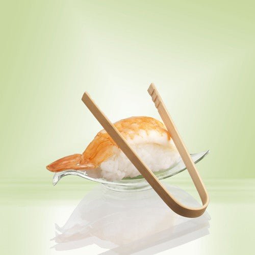 Pár 10 cm PAPSTAR kleští na finger food z bambusu od PAPSTAR GmbH spočívá na skleněné misce vedle nigiri s krevetami - ideální pro servírování občerstvení nebo párty finger food - před pozadím s bledě zeleným přechodem.