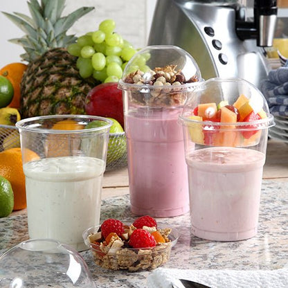 Tři smoothie v kelímcích na studené nápoje PLA "pure" průhledných 0,5 l od PAPSTAR GmbH stojí na kuchyňské lince. Dva mají ovoce a müsli pod vypouklými víčky. V pozadí jsou vidět čerstvé ovoce a mixér. Balení obsahuje 60 kusů.
