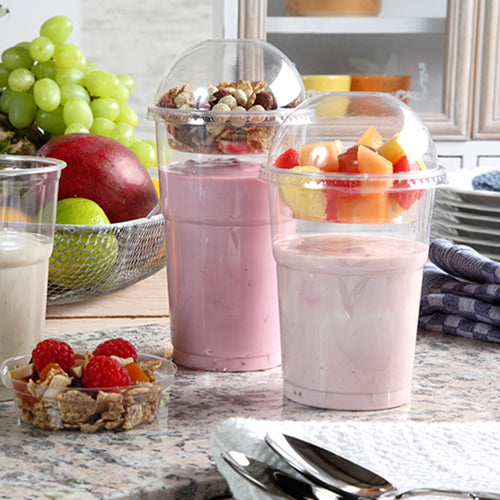 Dva jogurtové parfaity v plastových kelímcích, každý uzavřený Starpak 50 Dom-víčkem „Hurricane“ PET kulaté čiré Ø 9,5 cm, stojí na kuchyňské lince – jeden s müsli a ořechy, druhý s ovocem. V pozadí jsou vidět čerstvé bobule a naskládané talíře.