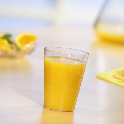 Jeden Starpak 10 kelímek na pití, PS 0,2l průhledný, naplněný pomerančovým džusem stojí na světlém stole. V pozadí jsou rozmazané plátky pomeranče a džbán.