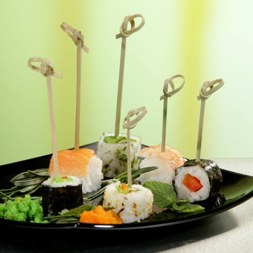 Je prezentován černý talíř s různými druhy sushi, které jsou napíchnuté na PAPSTAR 250 Fingerfood-Picker „pure“ od PAPSTAR GmbH. Celé je to ozdobeno jikrami, listovou zeleninou a wasabi; pozadí je v jemných zelených a žlutých tónech.