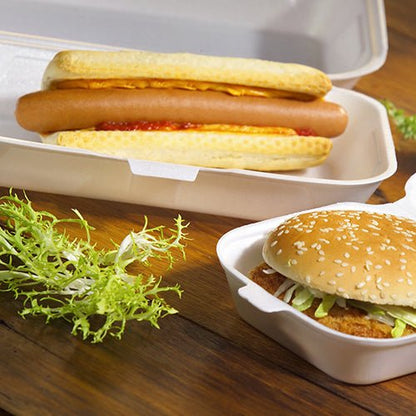 Hotdog s kečupem a hořčicí je servírován v univerzální krabici Starpak 100 s klopovým víkem XPS 1800 ml, vedle je sezamový hamburger se salátem v další krabici Starpak. Na dřevěném stole leží kadeřavý list salátu.
