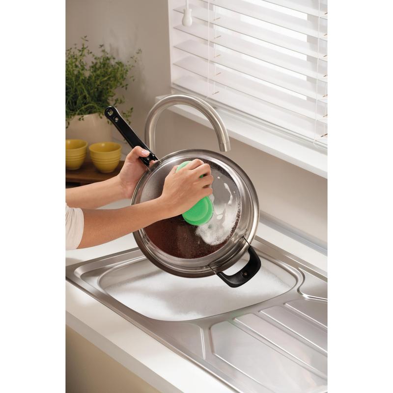 Někdo čistí kovovou pokličku nad mýdlovým dřezem s čisticí houbičkou Scotch-Brite® Classic od 3M Deutschland GmbH, v blízkosti okna s bílými žaluziemi a pultu s rostlinou a naskládanými žlutými miskami.