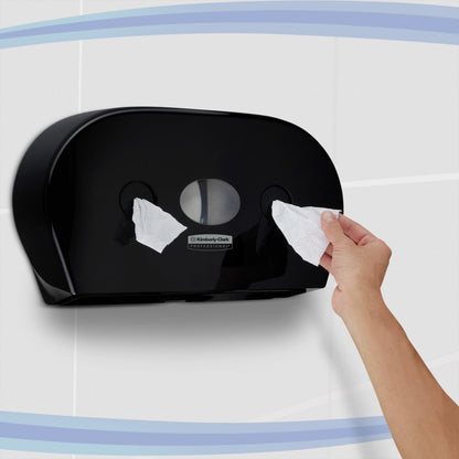 Osoba vytahuje toaletní papír z černého dávkovače toaletního papíru Kimberly-Clark Professional™ - centrální odběr (Jumbo) od společnosti Kimberly-Clark GmbH, který je připevněn na obložené zdi s modrými křivkovými pruhy.