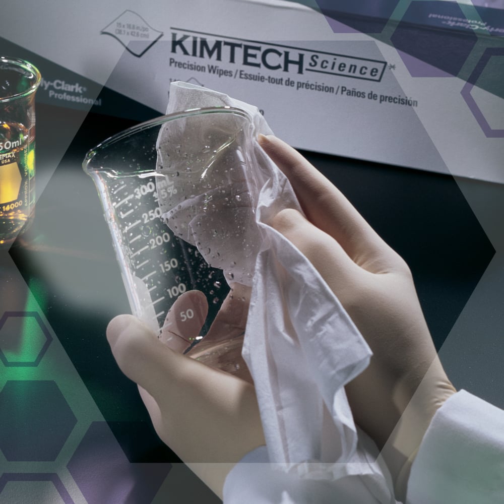 Osoba v bílých rukavicích utírá skleněný pohár s Kimtech® Science Precision ubrousky od Kimberly-Clark GmbH. V pozadí je krabice s ubrousky (100 listů, dvouvrstvé) a pohár s kapalinou.
