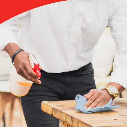 Osoba v bílém tričku s hodinkami čistí dřevěný stůl modrou utěrkou, možná WypAll® L20 General Clean - BRAG™ Box od společnosti Kimberly-Clark GmbH. Obrázek má nahoře červený zakřivený okraj, který zdůrazňuje důraz na detail.