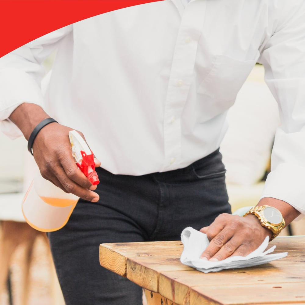 Osoba v bílé košili a černých kalhotách používá sprejovou láhev a roli WypAll® L40 PowerClean (Kimberly-Clark GmbH) k čištění dřevěného stolu. Osoba nosí hodinky; horní okraj obrázku ukazuje červený zakřivený okraj.