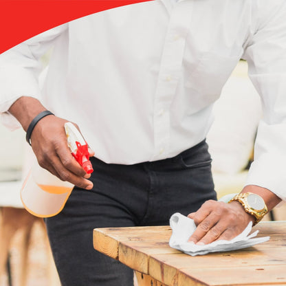 Osoba v bílé košili a černých kalhotách používá sprejovou láhev a roli WypAll® L40 PowerClean (Kimberly-Clark GmbH) k čištění dřevěného stolu. Osoba nosí hodinky; horní okraj obrázku ukazuje červený zakřivený okraj.