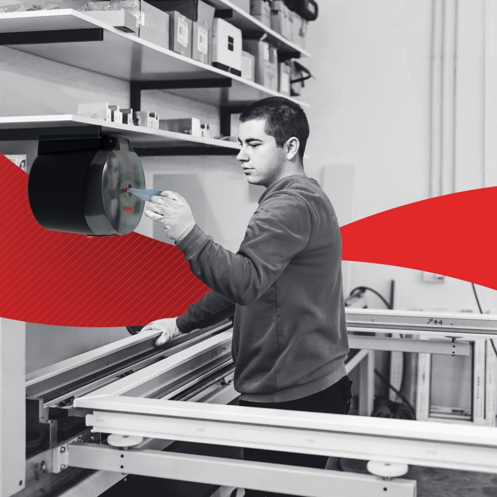 V dílně obsluhuje osoba zařízení namontované na zdi poblíž regálů s WypAll® L10 papírovými utěrkami pro potraviny a hygienu Centrefeed (6 rolí) od Kimberly-Clark GmbH; nad tím jsou krabice a červený pruh zvýrazňuje pozadí.