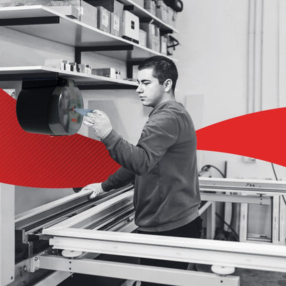 V dílně obsluhuje osoba zařízení namontované na zdi poblíž regálů s WypAll® L10 papírovými utěrkami pro potraviny a hygienu Centrefeed (6 rolí) od Kimberly-Clark GmbH; nad tím jsou krabice a červený pruh zvýrazňuje pozadí.