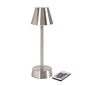 LED lampa bezdrátová ZELDA stříbrná od Duni GmbH s kuželovým stínidlem a válcovou základnou stojí na bílém pozadí. Vedle ní leží dálkové ovládání pro energeticky úsporná, moderní osvětlovací řešení (1 kus).