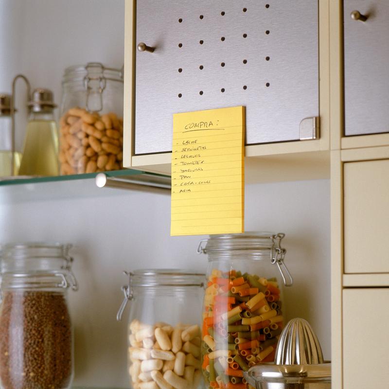 Ručně psaný nákupní seznam ve španělštině se nachází na béžové kuchyňské skříňce s použitím 3M Deutschland GmbH Post-it® Super Sticky Notes ve velkém formátu, Ultra Žlutá (101 x 152 mm), nad sklenicemi s těstovinami, fazolemi a obilovinami. V sousedních policích jsou další sklenice a kuchyňské náčiní.