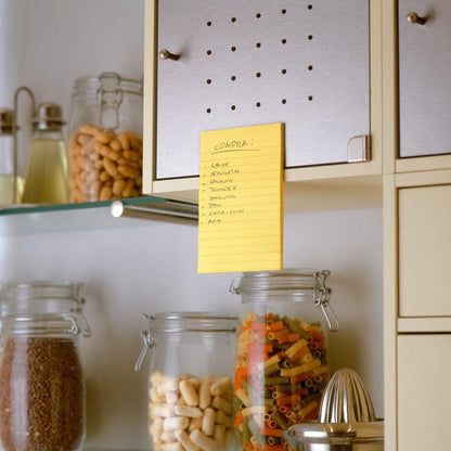 Ručně psaný nákupní seznam ve španělštině se nachází na béžové kuchyňské skříňce s použitím 3M Deutschland GmbH Post-it® Super Sticky Notes ve velkém formátu, Ultra Žlutá (101 x 152 mm), nad sklenicemi s těstovinami, fazolemi a obilovinami. V sousedních policích jsou další sklenice a kuchyňské náčiní.