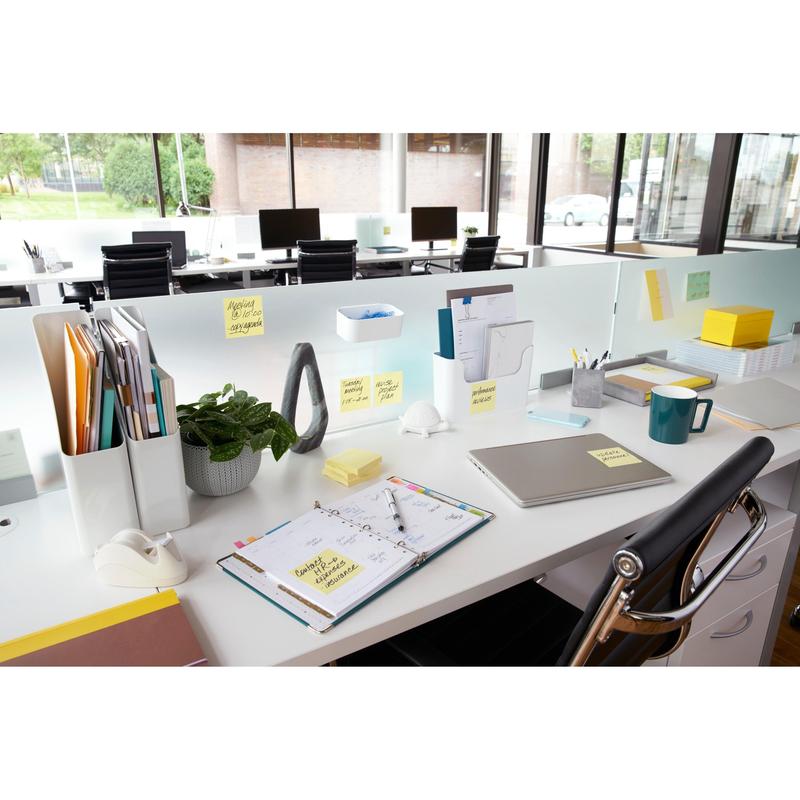 Moderní kancelářský stůl s laptopem, plánovačem, hrnkem na kávu, rostlinou, psacími potřebami a žlutými Post-it® Notes od 3M (76x76 mm, 100 listů/blok), ideální pro rychlé poznámky. Dokumenty jsou uspořádány a velká okna poskytují výhled ven; stoly jsou uspořádány v řadě.