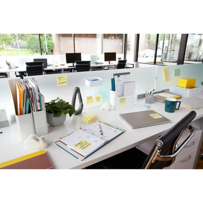 Moderní kancelářský stůl s laptopem, plánovačem, hrnkem na kávu, rostlinou, psacími potřebami a žlutými Post-it® Notes od 3M (76x76 mm, 100 listů/blok), ideální pro rychlé poznámky. Dokumenty jsou uspořádány a velká okna poskytují výhled ven; stoly jsou uspořádány v řadě.