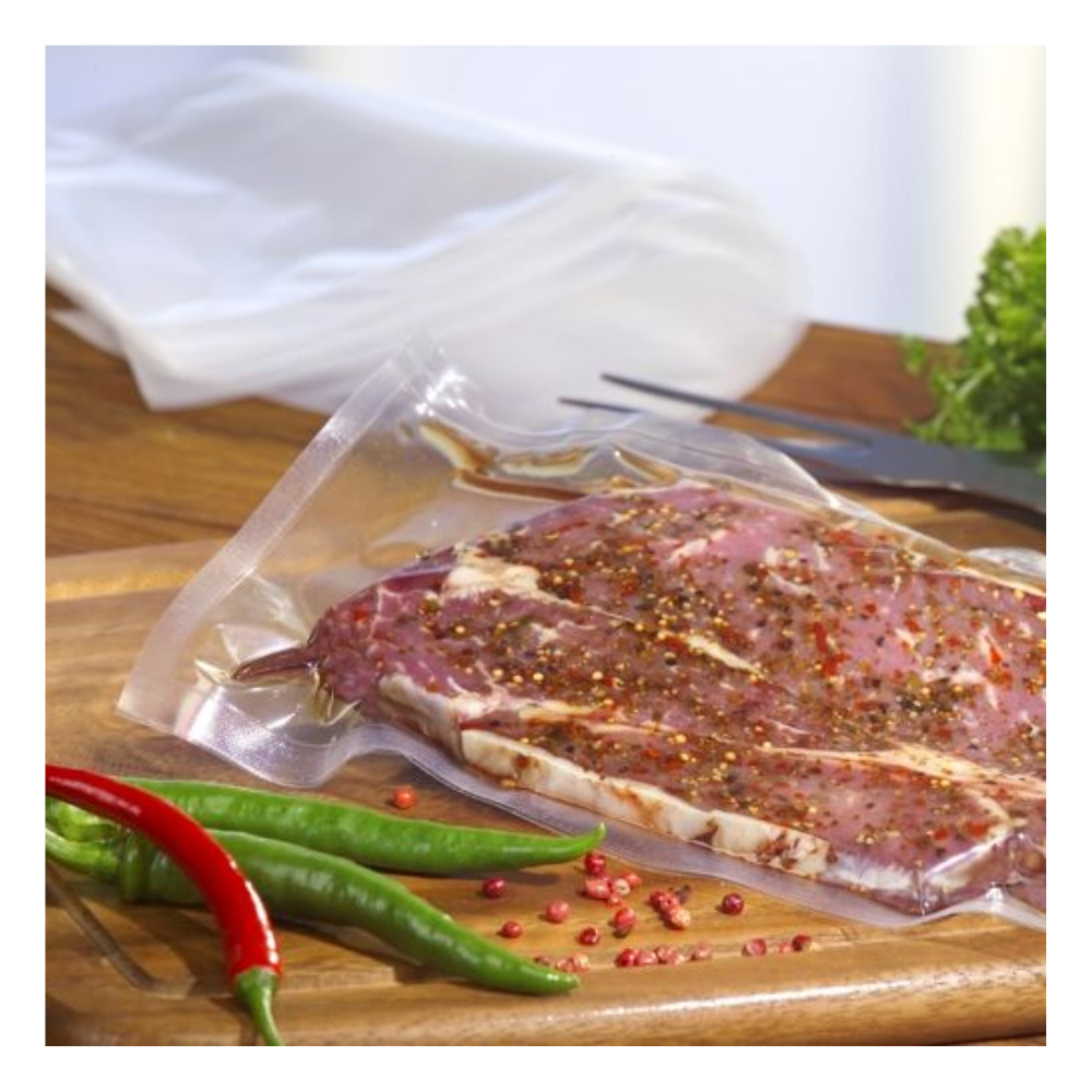 Syrový, marinovaný steak je vakuován v průhledném sáčku Starpak 100 (PA/PE) s komorovým vakuovačem na dřevěném prkénku, s chilli papričkami, růžovými pepřovými zrny, listovou zeleninou a vidličkou v pozadí.