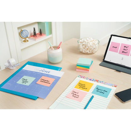 Organizovaný stůl pro učení nebo práci s Post-it® Notes (76 mm x 127 mm, 100 listů/blok, 6 bloků/balení, 100% PEFC) od 3M Deutschland GmbH, poznámkovými bloky, pery v hrnku, popcornem, lepicí páskou, tabletem s poznámkami; knihovna s knihami zdobí pozadí.