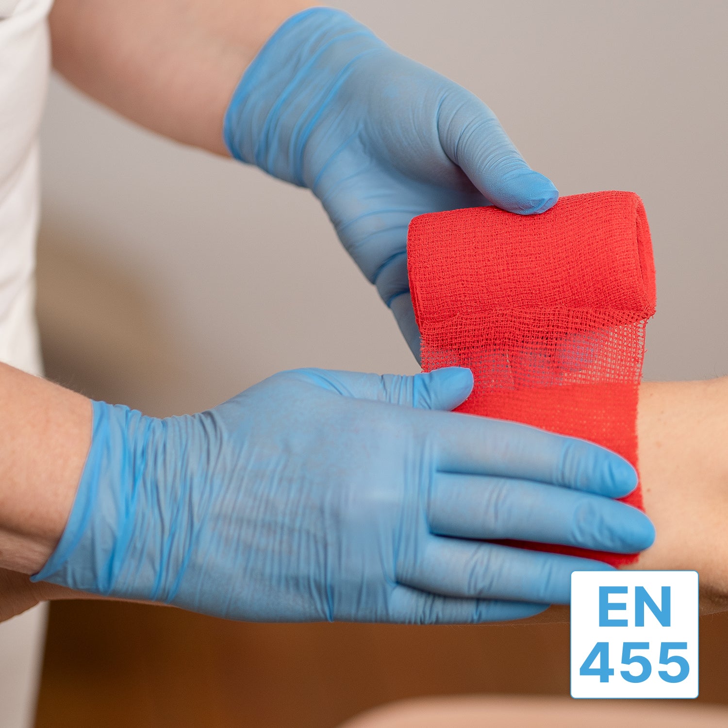 Eine Person, die ARNOMED NITRIL BLUE 200er Nitrilhandschuhe Blau von ARNOWA GmbH trägt, wickelt eine rote Bandage um den Unterarm einer Person. Ein kleines blau-weißes Etikett mit der Aufschrift "EN 455" erscheint in der unteren rechten Ecke des Bildes.