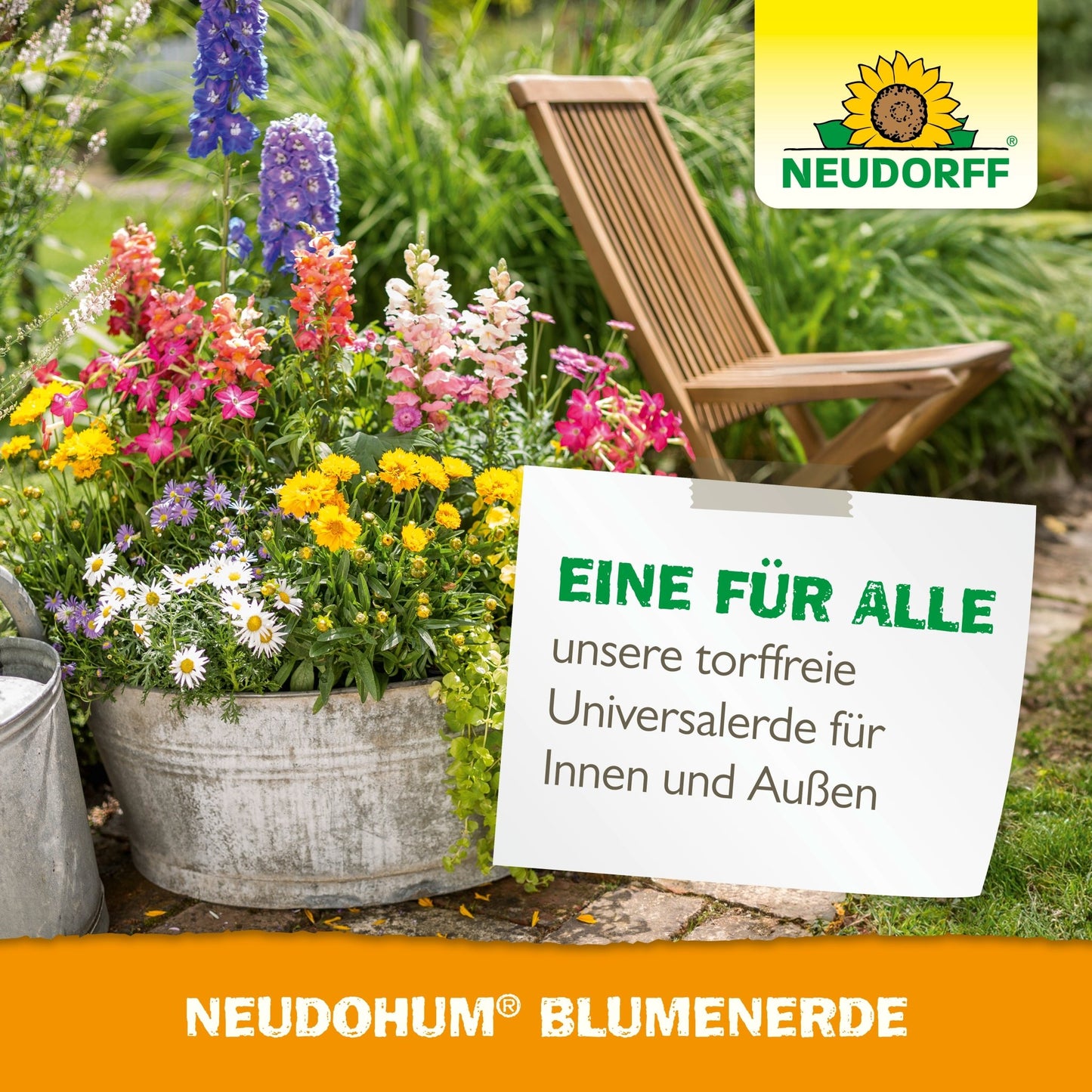 Kovový kýbl s barevnými zahradními rostlinami stojí na zahradní cestě vedle dřevěné skládací židle. Cedule propaguje bezrašelinovou NeudoHum BlumenErde od Neudorff Shop pro vnitřní a venkovní použití.