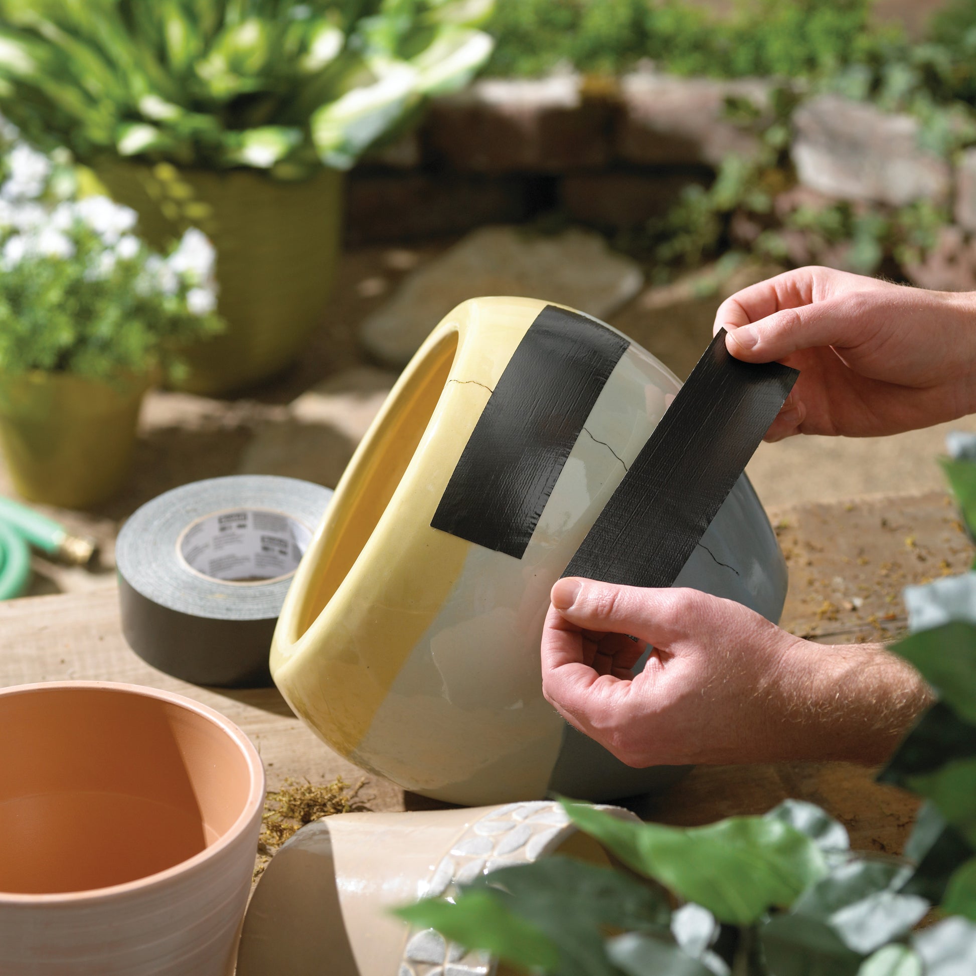 Eine Person klebt 3M Scotch® Extremium™ All Weather Hochleistungs-Klebeband (27 m x 48 mm) auf einen zerbrochenen Keramiktopf auf Holz. Im Hintergrund sind weitere Töpfe, Klebeband, Grünpflanzen und Gartengeräte zu sehen.
