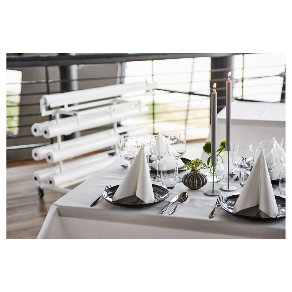 Na hezky prostřeném jídelním stole stojí čerstvá, bílá Duni Dunicel® ubrusová role od Duni GmbH, která nabízí látkově podobnou texturu. Trojúhelníkově složené ubrousky na tmavých talířích doprovázejí sklenice na víno. Vysoké svíčky a malé květinové aranžmá slouží jako stolní dekorace, v pozadí jsou vidět srolovaná plátna.