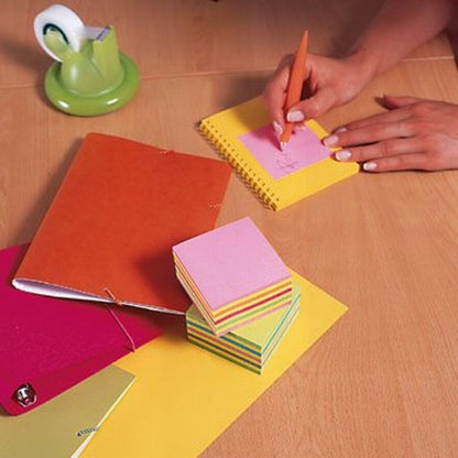 An einem hölzernen Schreibtisch schreibt jemand mit einem orangefarbenen Stift auf einen rosa Post-it® Notes Würfel (76 mm x 76 mm, 450 Blatt/Block, 100% PEFC) der 3M Deutschland GmbH. In der Nähe liegen ein rotes Notizbuch, ein grüner Klebebandabroller, eine silberne Glocke und weitere bunte Zettel.