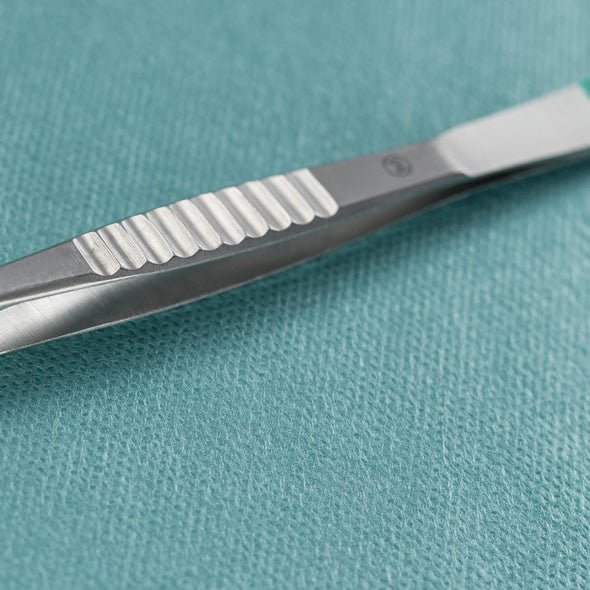 Detailní záběr na Hartmannovu štěpící pinzetu Paul Hartmann AG rovnou 9 cm, sterilní (balení po 40 kusech), ležící vedle chirurgického skalpelu z kovu na tyrkysově zbarvené, strukturované lékařské látce.