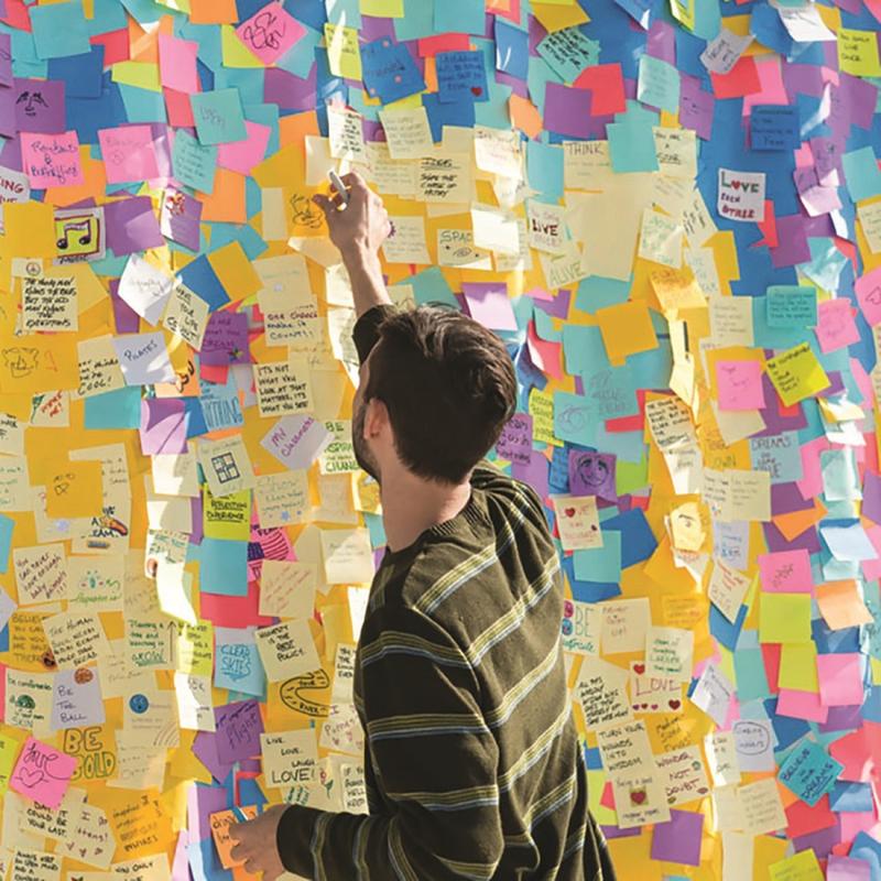 Osoba v pruhovaném tričku stojí u stěny s ručně psanými zprávami a kresbami a sahá po kostce Post-it® Notes v ultra barvách od 3M Deutschland GmbH (76x76 mm, 450 listů, 100% PEFC), aby ji umístila mezi barevné lístky.