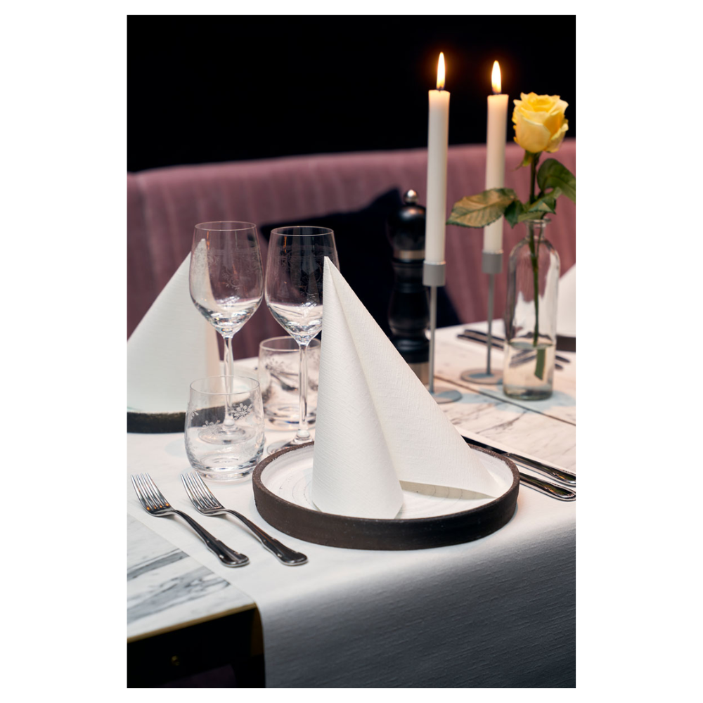 Jídelní stůl je elegantně prostřený dvěma ubrousky Duni Dunilin® 48 x 48 cm Bílý, složenými do trojúhelníků na talířích. Dvě velké hořící svíčky a žlutá růže ve skleněné váze zdobí pozadí. Sklenice na víno, sklenice na vodu a příbory doplňují toto sofistikované prostředí s látkovým šarmem.
