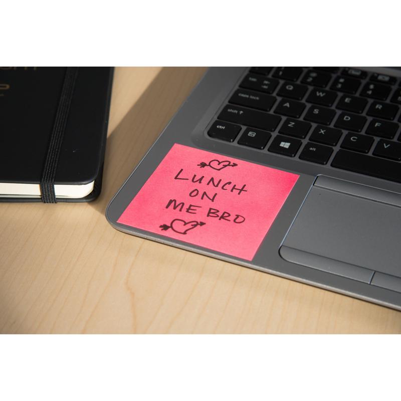 Růžový Post-it® Super Sticky Z-Note (76x76mm) od 3M Deutschland GmbH s nápisem "Lunch on me bro" černým inkoustem a dvěma srdíčky leží na klávesnici laptopu vedle černého notebooku, vše na světlém dřevěném stole.