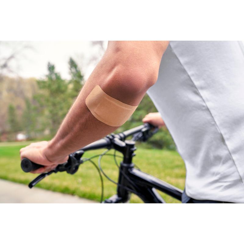 Osoba, která má na sobě bílé tričko a jezdí venku na kole, má na holé paži náplast Nexcare™ Ultra Stretch MAXI Comfort Flexible (50 mm x 101 mm) od 3M Deutschland GmbH; v pozadí je vidět zelená tráva a stromy.