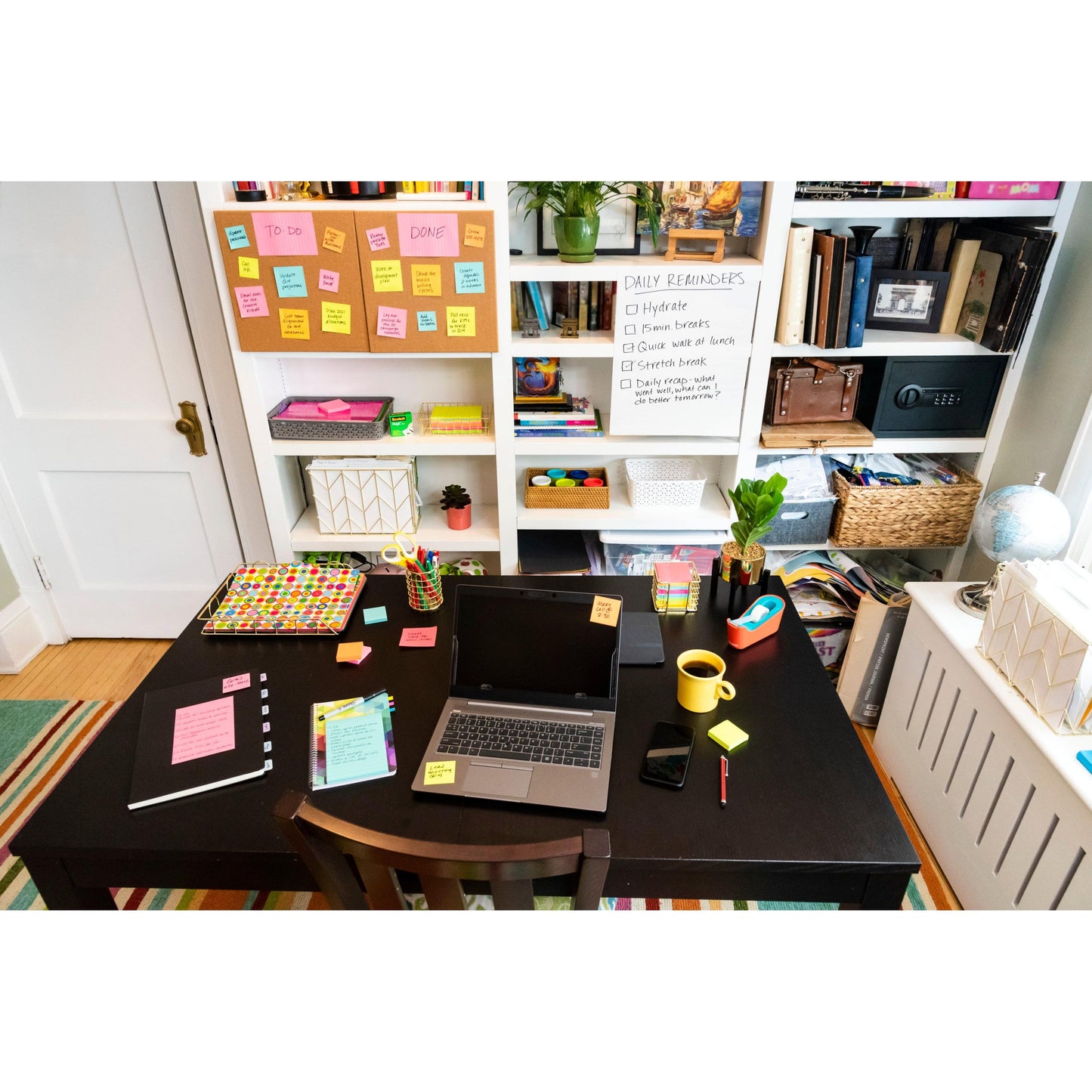 Ein Schreibtisch mit Laptop, Notebook, Telefon und 3M Deutschland GmbH Post-it® Super Sticky Notes (76 mm x 76 mm, 90 Blatt/Block, 100% PEFC) steht vor einem Bücherregal mit Büchern und einem Whiteboard für die täglichen Erinnerungen; bunte Materialien sorgen für Ordnung.