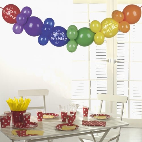 Bílý stůl s party nádobím s červenými puntíky, žlutými příbory a kelímky je korunován girlandou z balónků 2 m „Happy Birthday“ od PAPSTAR GmbH - slavnostní narozeninová dekorace, která se ve slunečním světle třpytí skrze bílé okenice.