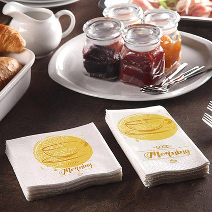 Snídaňový stůl s croissanty, konvicí a 200 PAPSTAR ubrousky (3-vrstvé 1/4-skládané, 24x24 cm) od PAPSTAR GmbH s nápisem „Dobré ráno“, k tomu sklenice s marmeládou a tři vidličky pro pohodovou snídani.