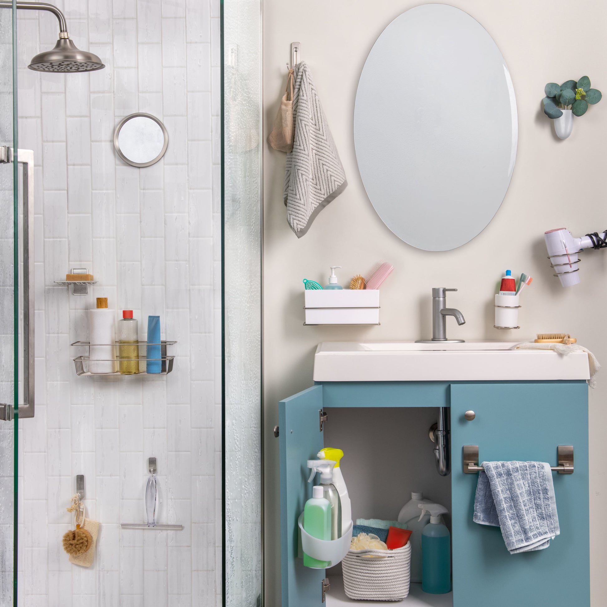 Ein Badezimmer mit einer Glasdusche auf der linken Seite, geordneten Regalen, einem Waschbecken mit blauem Schrank darunter, in dem Reinigungsmittel aufbewahrt werden, einem ovalen Spiegel darüber und einem 3M Command™ Großer Doppelhaken von 3M Deutschland GmbH, der ein Handtuch am Schrankgriff hält.