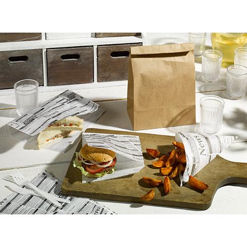 Na stole pokrytém balicím papírem, pergamenová náhrada 35x25 cm odolná proti mastnotě od PAPSTAR GmbH, je servírován sendvič, burger a bramborové klínky, vedle jsou průhledné poháry, hnědý papírový sáček, džbán se žlutým nápojem a bílé plastové příbory.