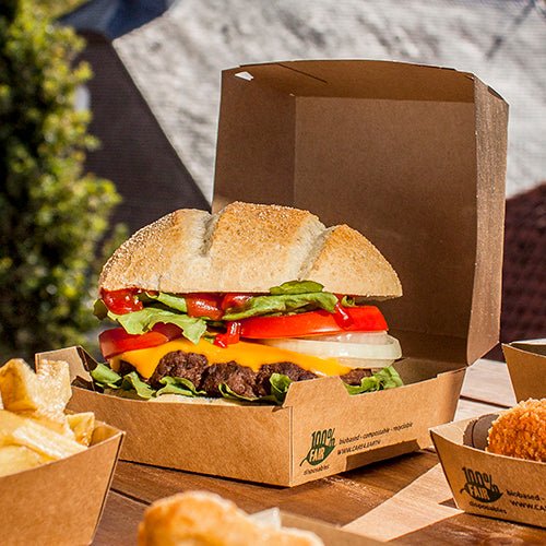 Cheeseburger se salátem, rajčetem, cibulí a čedarem leží venku v PAPSTAR GmbH burgerové krabici z kartonu (9x18,5x18,5 cm, "100% Fair", 25 kusů). Hranolky a další přílohy jsou podávány samostatně vedle.