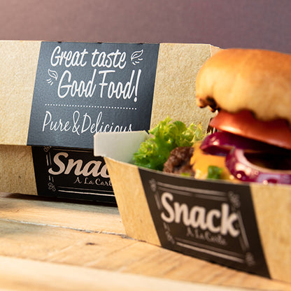 Papírový tácek s nápisem "Snack A La Carte" obsahuje hamburger se salátem, rajčetem a cibulí. V pozadí stojí PAPSTAR 50 burgerových krabic, karton "pure" "Good Food" od PAPSTAR GmbH: Dobrý chuť, Good Food! Čisté & lahodné.