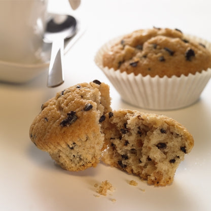 V popředí je vidět polovina čokoládového muffinového čipu, za ní celý muffin v PAPSTAR 1000 kulatých pečicích košíčků od PAPSTAR GmbH. V pozadí jsou nakrojený bílý šálek s podšálkem a lžičkou.