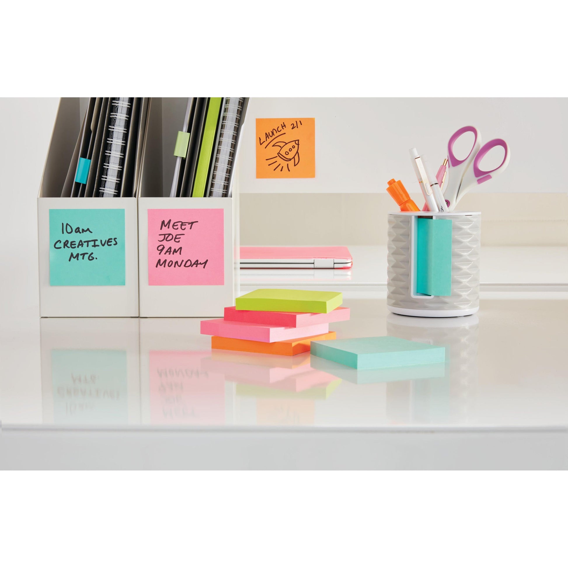 Ein Schreibtisch mit 3M Deutschland GmbH Post-it® Notes, Energetic Collection (38 mm x 51 mm), zwei weißen Zeitschriftenhaltern, einem Stifthalter mit Stiften und Scheren sowie Notizbüchern. Die Haftnotizen zeigen Erinnerungen und Kritzeleien.