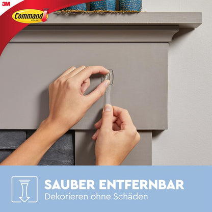 Osoba stahuje průhledný lepicí proužek z šedého krbu a ukazuje, jak Command™ Mini-Háčky, průhledné, s průhlednými proužky (17006CLR) od 3M Deutschland GmbH lze bez stopy dekorovat a připevnit. Loga Command a 3M jsou viditelná.