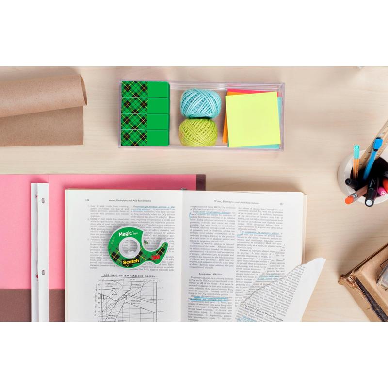 Stůl s otevřenou knihou, šanony, pastelky v držáku, samolepicí poznámky, dvě koule provázku, několik zelených gum, role hnědého papíru a balení neviditelné lepicí pásky 3M Scotch® Magic™ (8 rolí, 19 mm x 33 m).