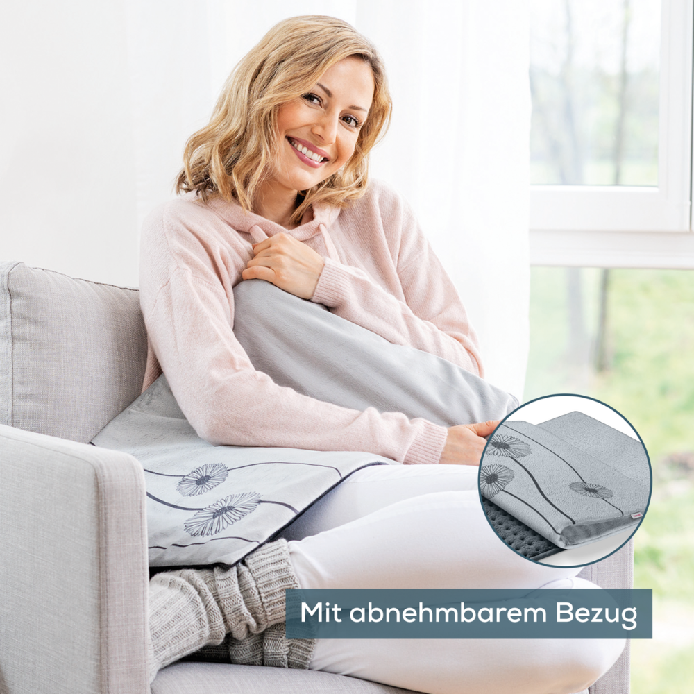 Žena sedí na světle šedé pohovce vedle okna a drží Beurer HK 125 XXL vyhřívací polštář Cosy Grey od Beurer GmbH v ruce. Usmívá se a má na sobě růžovou mikinu s kapucí. Je zde detailní záběr polštáře, který ukazuje jeho odnímatelný potah a topné funkce se 6 úrovněmi tepla. Německý text zní „S odnímatelným potahem.“