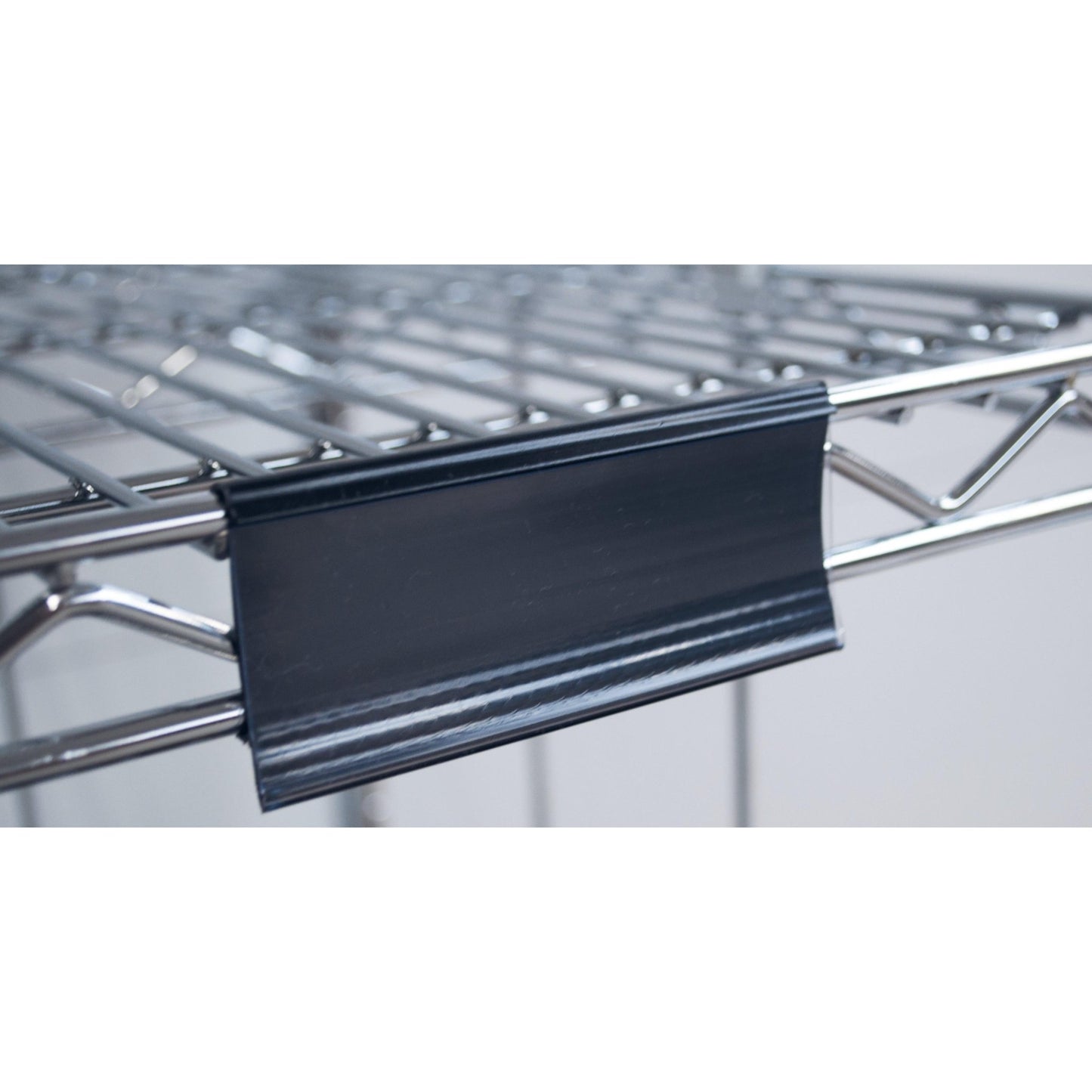 Detailní záběr na rámeček štítku Novocal s papírovými štítky (cca 100 x 44 mm, 1 kus), připevněný na kovovém drátěném regálu, s černým plastovým držákem štítku na předním okraji; pozadí je rozmazané.