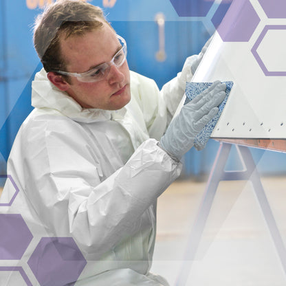 Eine Person in Schutzkleidung benutzt eine Kimtech® Prozeßwischtücher Großrolle (Kimberly-Clark GmbH), um einen großen weißen Gegenstand in einer industriellen Umgebung zu reinigen. Eine violette Hexagon-Grafik überlagert das Bild.