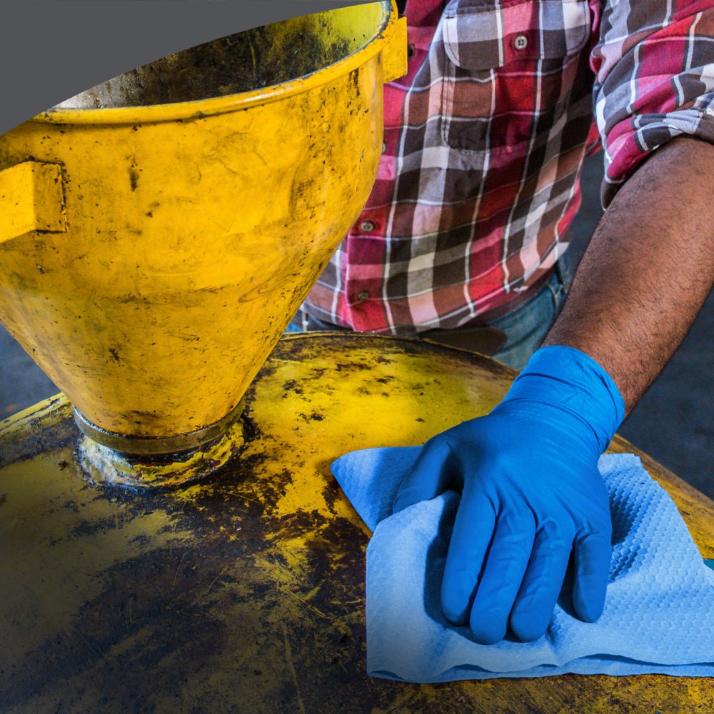 Osoba v kostkované košili a džínách otírá žlutý průmyslový stroj modrým WypAll® X70 PowerClean čisticím ubrouskem od Kimberly-Clark GmbH ze Zupfboxu, čímž zajišťuje profesionální úklid.