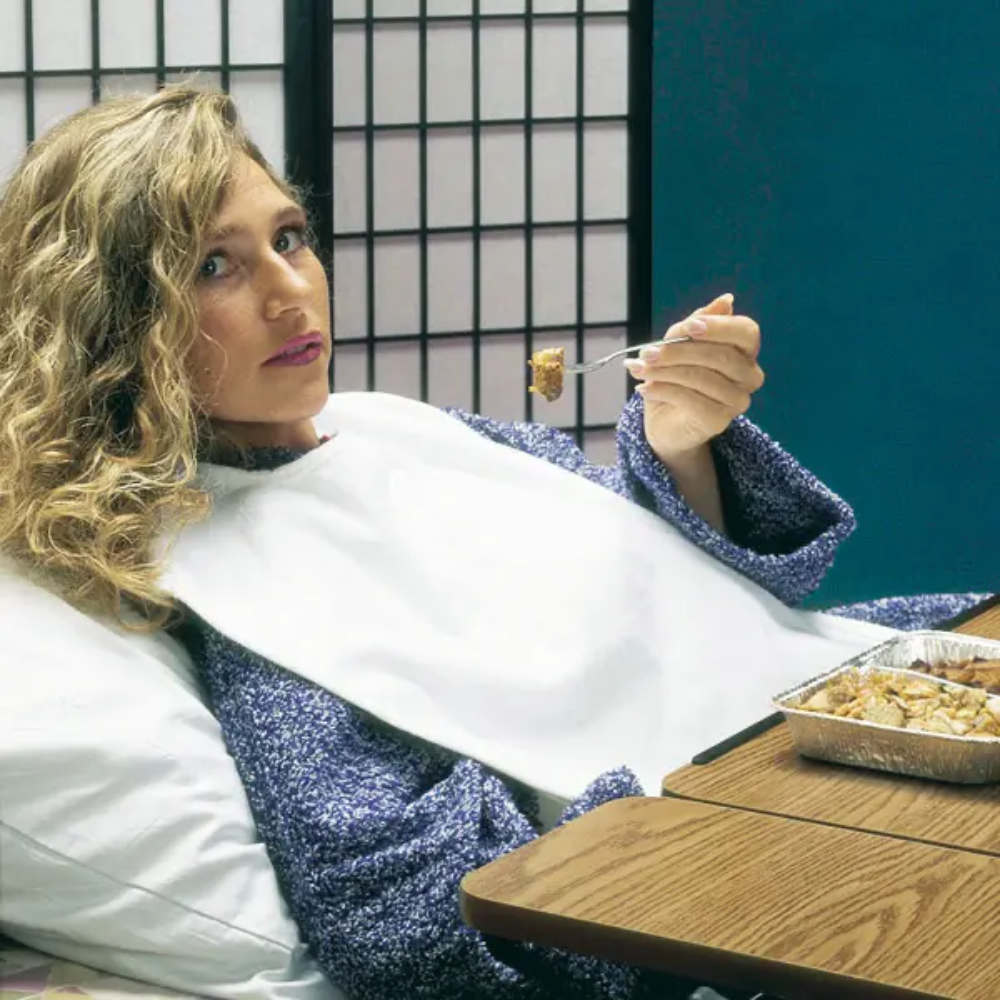 Osoba s kudrnatými vlasy leží v posteli opřená o polštář, má na sobě modrý svetr a ochranu oděvu pro dospělé Servocare od společnosti Servoprax GmbH. Jí vidličkou jídlo z fóliového obalu. Před ní stojí dřevěný stůl a v pozadí je shoji paraván.