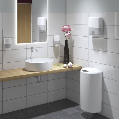 Ein modernes Badezimmer präsentiert ein rundes weißes Waschbecken auf einer Holzplatte, geschmückt mit einer hohen Vase mit rosa Blumen. Der Tork 561000 Mini Spender für Flüssigseife Elevation S2 von TORK – Essity Professional Hygiene Germany GmbH sorgt unter dem großen beleuchteten Spiegel für Funktion und Stil. Seifenspender an den weiß gefliesten Wänden unterstreichen das Elegance Design, während ein eleganter weißer Mülleimer auf den kontrastierenden dunklen Bodenfliesen steht.
